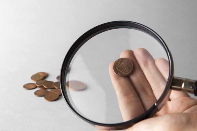 A coin under a magnifying glass and other coins on the table in the concept of 'The Hidden Costs in Home Removals Services'.
